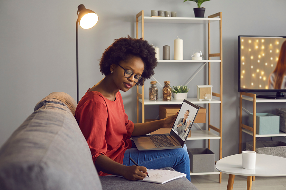 Woman watches a video presentation on her laptop while sitting on a couch taking notes